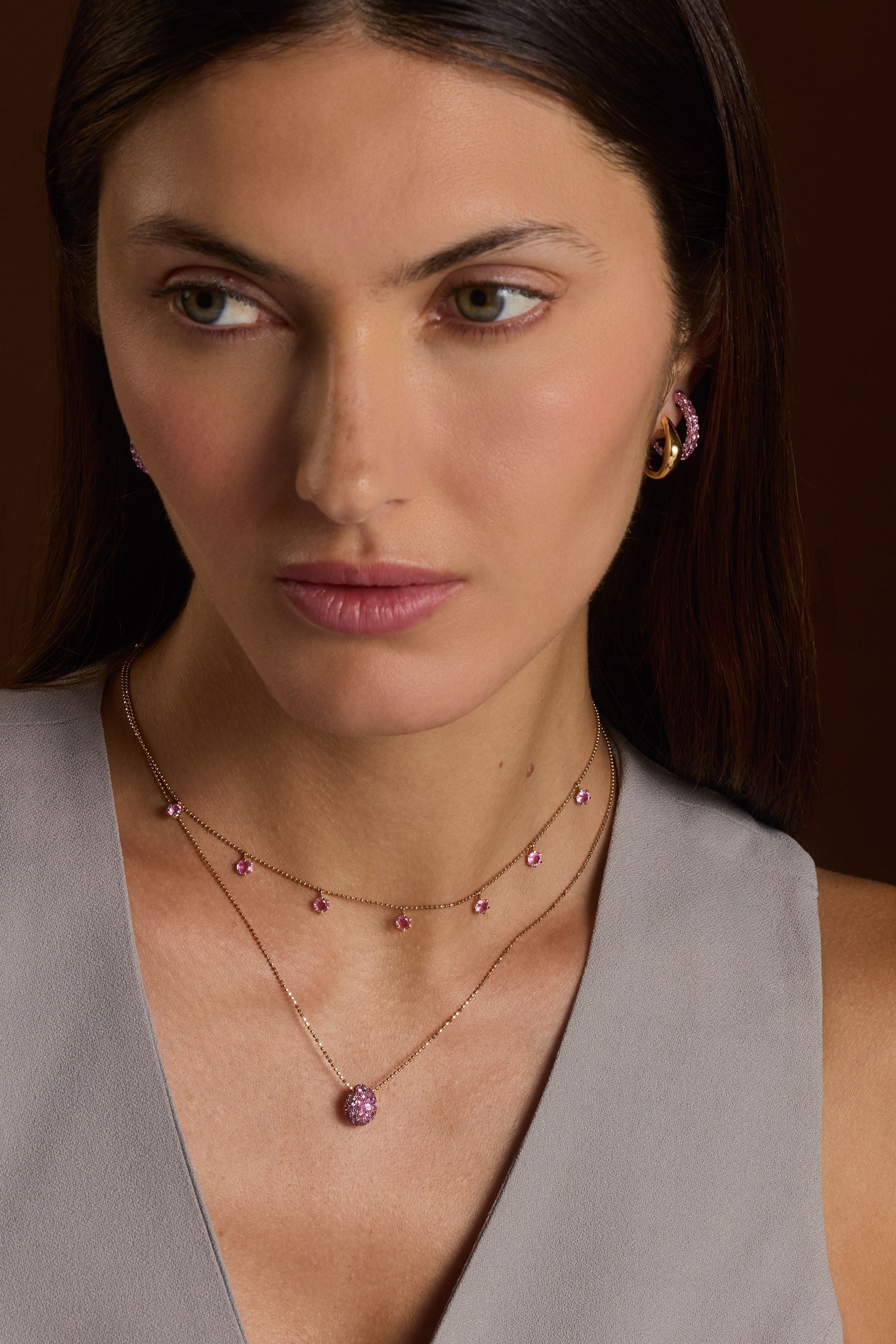 Woman wearing pink sapphire floating necklace and three sided hoop earrings against a brown background